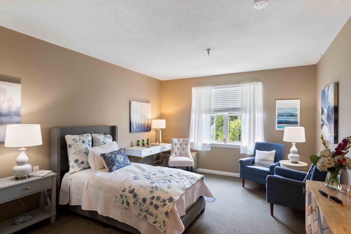 A cozy bedroom with a well-decorated interior featuring a bed, chairs, and natural light coming through the window.