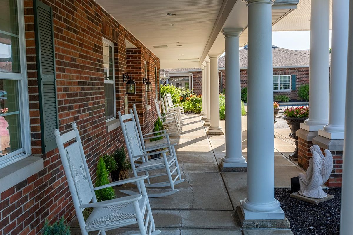 A sunlit porch with white rocking chairs lined up outside a brick building.