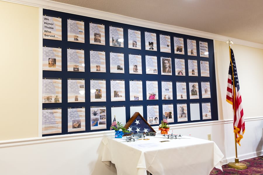 A memorial wall displays framed certificates and tributes to military veterans, with a folded flag and flowers on the table below.