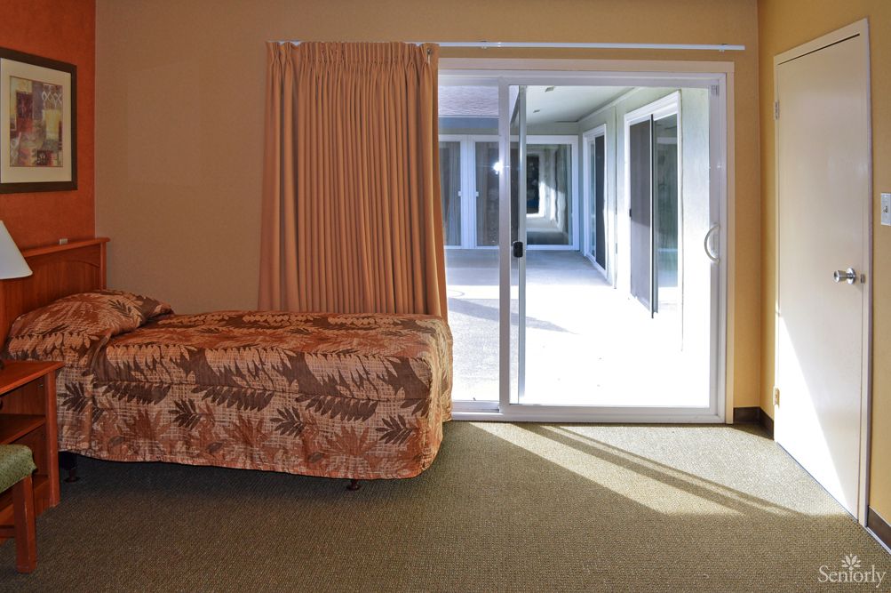 A cozy bedroom featuring a bed with a floral bedspread, adjacent to a sliding glass door leading to a sunlit patio.