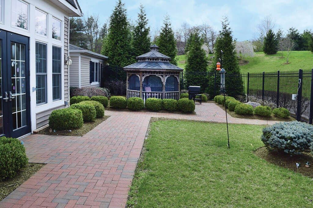 A well-maintained garden features a brick pathway leading to a gazebo, surrounded by neatly trimmed shrubs and lush green trees.