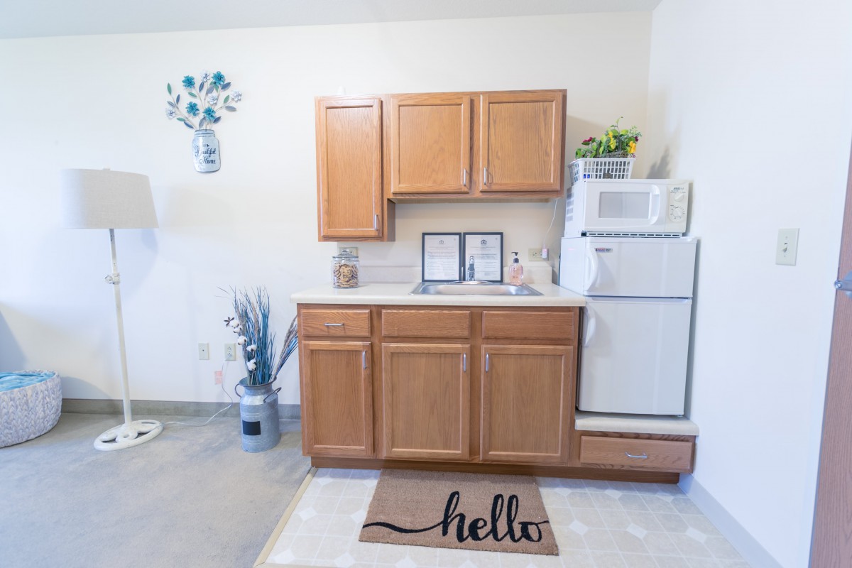 A compact kitchen setup with wooden cabinets, a small fridge, decorative plants, and a welcoming doormat.