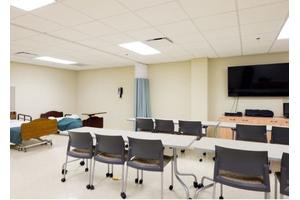 A hybrid classroom and medical training room with desks, chairs, a hospital bed, and a television.