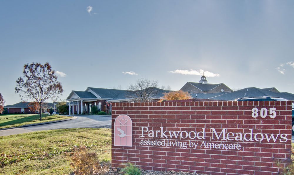 The image shows the entrance of Parkwood Meadows Residential Living, featuring a brick sign and part of the building in the background under a clear sky.