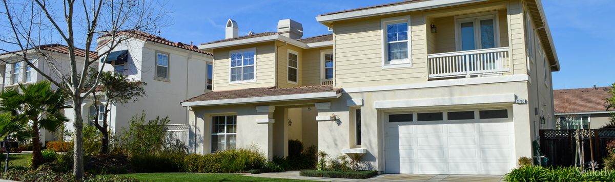 A suburban two-story house with beige siding and white trim under a clear blue sky.