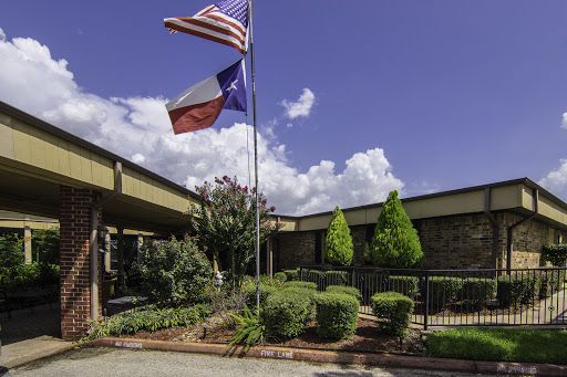 A view of a building's entrance with American and Texas flags flying, surrounded by well-maintained landscaping under a clear blue sky.