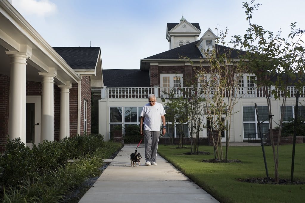 A man walks his dog along a sidewalk in front of a large house.