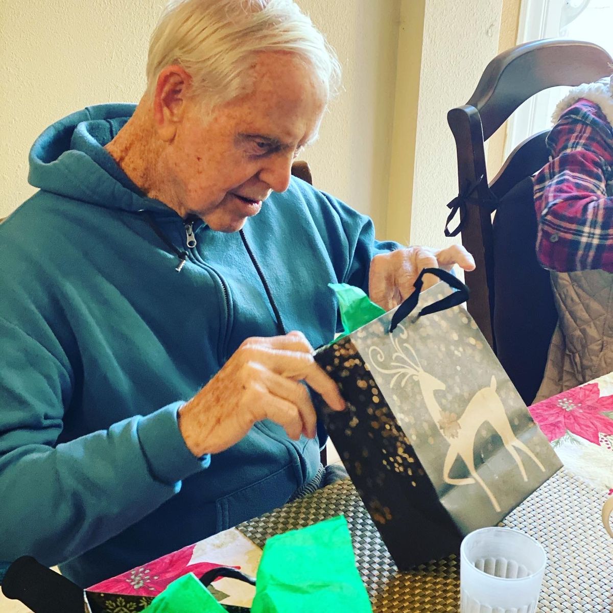 An elderly man in a hoodie is opening a gift at a table indoors.