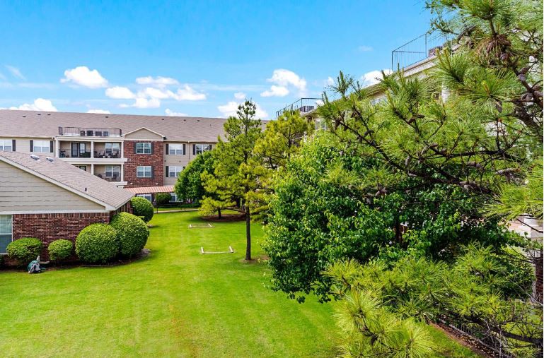A well-maintained green space with trees and a lawn surrounded by an apartment complex under a clear blue sky.