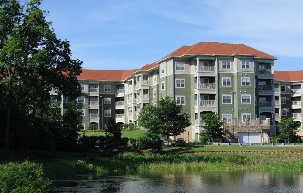 A residential apartment building with a green facade and red roof, situated near a pond and surrounded by trees.