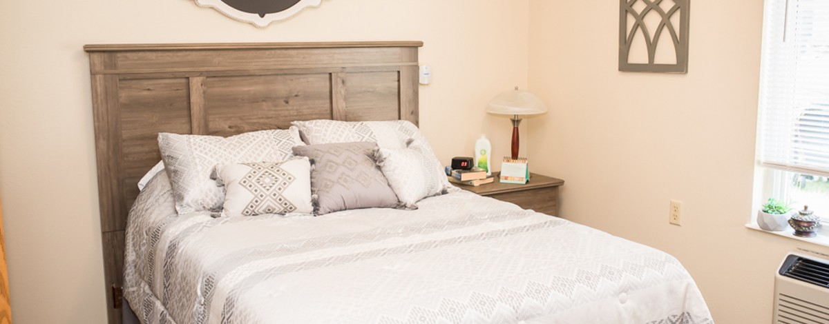 A cozy bedroom with a neatly made bed, adorned with several pillows, and a nightstand featuring a lamp and some items.