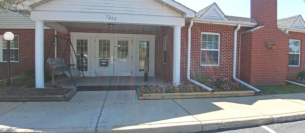 A brick building entrance with a double door and windows, featuring a small awning and adjacent flowerbed.