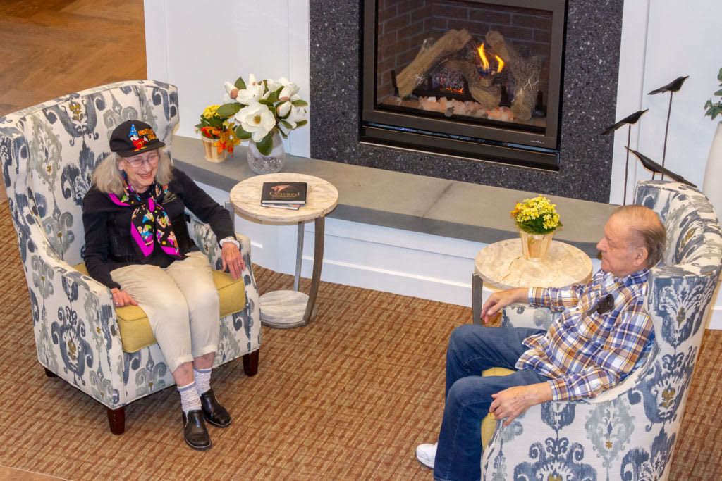 Two seniors seated by the fireplace engaging in conversation.