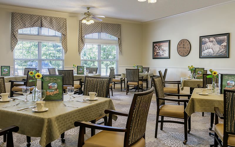 A cozy dining room with neatly arranged tables and chairs, featuring large windows and pleasant decor.