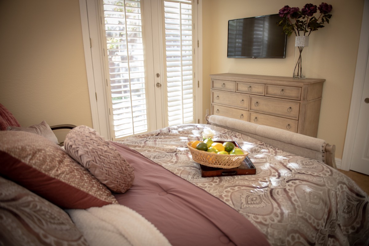 A cozy bedroom featuring a neatly made bed, a dresser with a television mounted above it, and natural light streaming through the shutters. A tray with fresh fruit adds a touch of hospitality to the space.