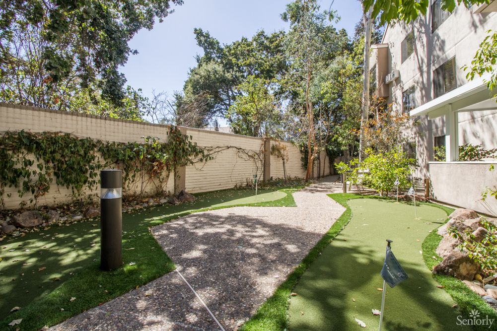 A serene garden area with a paved pathway, surrounded by trees and greenery, enclosed by a fence.
