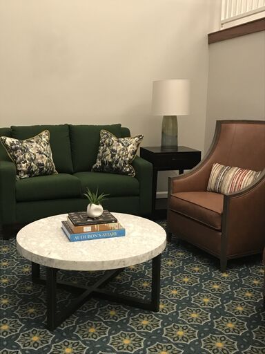 A cozy living room setup featuring a green sofa with patterned pillows, a brown armchair with a striped pillow, and a round marble coffee table topped with books and a decorative plant.