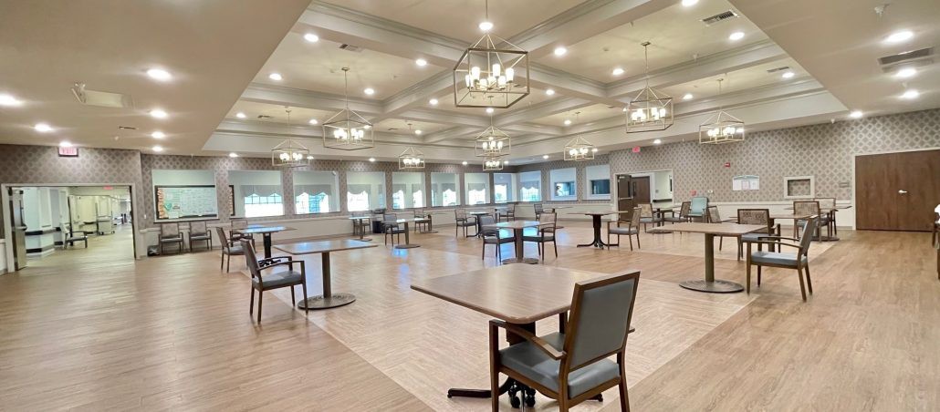 A spacious dining hall with multiple tables and chairs, illuminated by chandeliers and natural light from large windows.