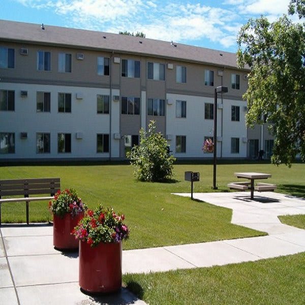 A sunny day view of an apartment building with a well-maintained lawn, flower pots, and benches along the pathway.