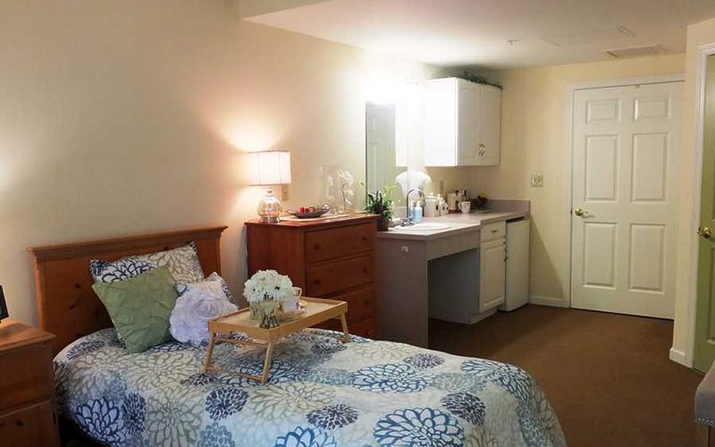 A cozy bedroom with a bed, dresser, and a small kitchenette area.