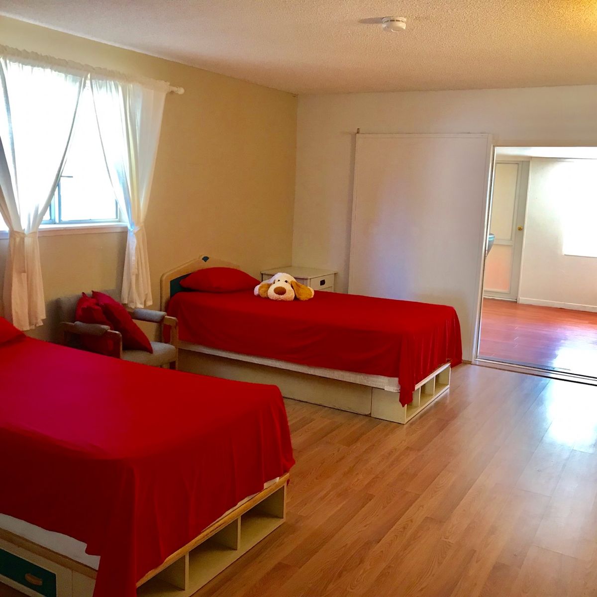 A spacious bedroom featuring two twin beds with bright red bedding, wooden floors, and natural light streaming through the window.