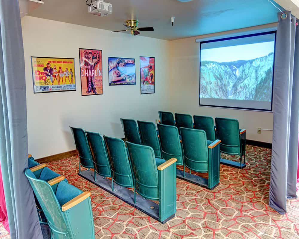 A small theater room with rows of green upholstered seats facing a large screen displaying a nature scene, adorned with colorful posters on the walls.