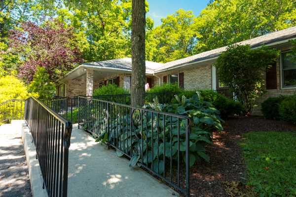 A charming single-story house surrounded by lush greenery and a paved walkway.