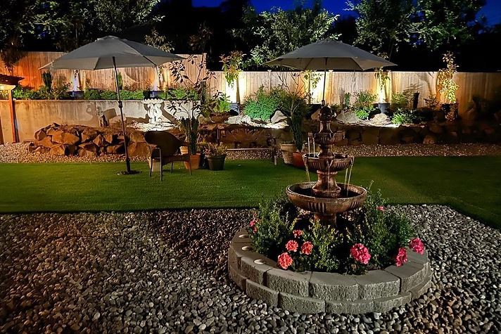 A beautifully lit garden at night featuring a central fountain, surrounded by flowers and outdoor seating areas.