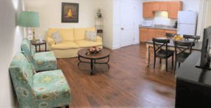 A cozy living room with a yellow sofa, coffee table, and adjacent kitchen area featuring wood flooring.