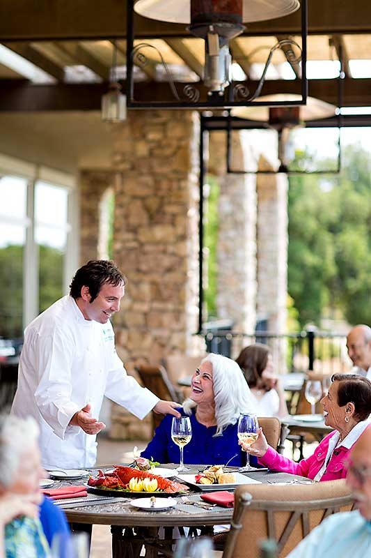 A chef engaging with guests at an outdoor restaurant table, with patrons enjoying wine and conversation.