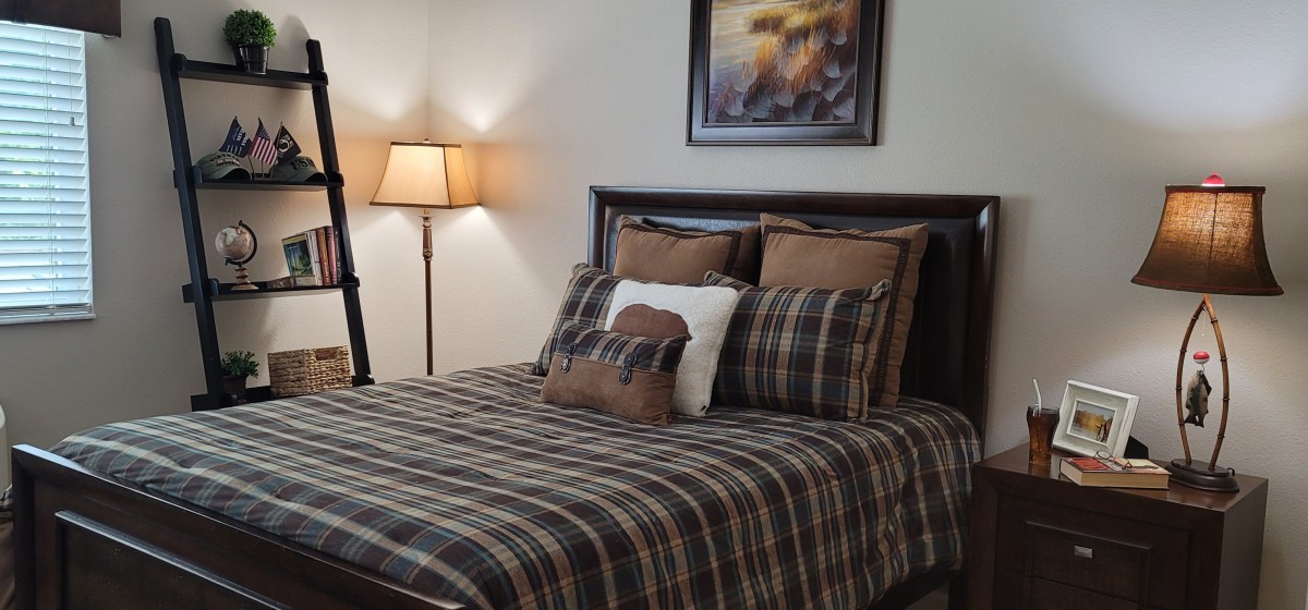 A cozy bedroom featuring a plaid-patterned bedspread and pillows, with a lamp on the side and framed artwork on the wall.