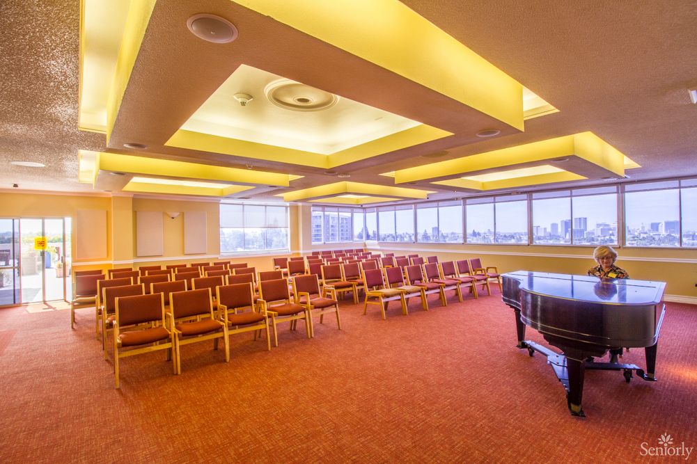 A spacious conference room with rows of seating, featuring a grand piano and large windows showcasing a cityscape.