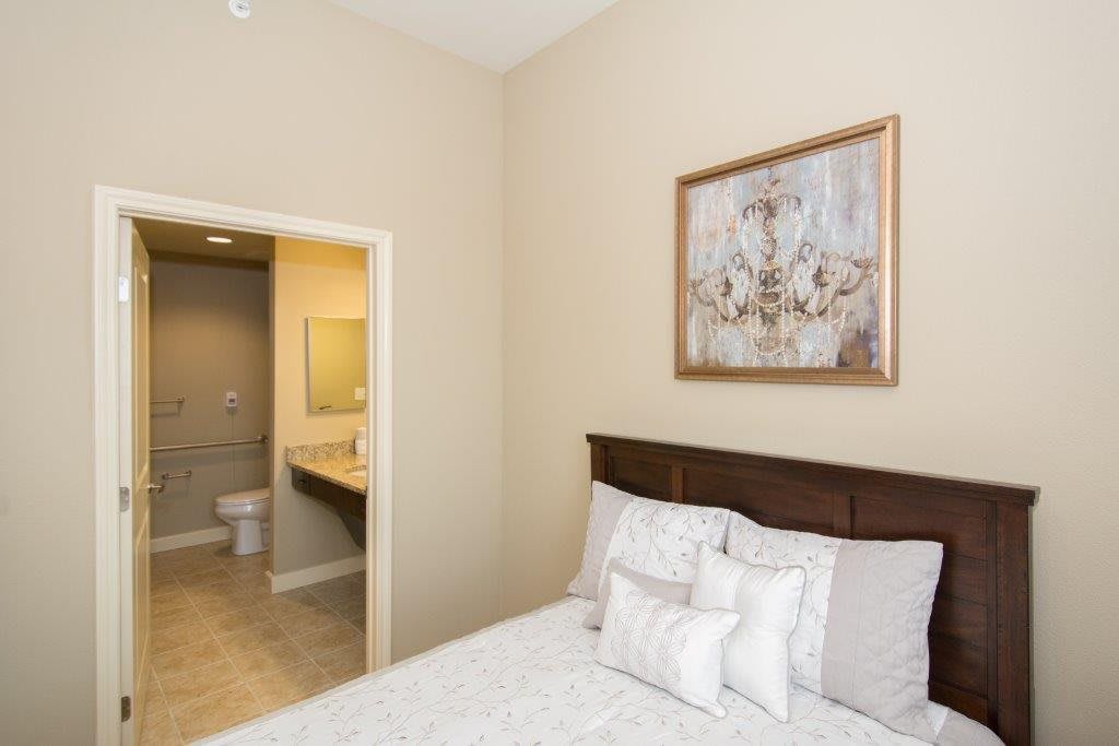A cozy bedroom with an adjacent bathroom, featuring a bed with white bedding and a decorative painting on the wall.