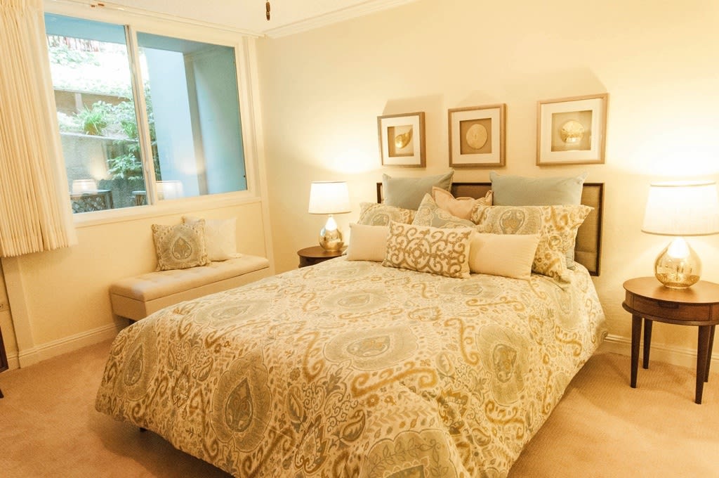 A cozy and well-decorated bedroom featuring a comfortable bed with patterned bedding and several pillows, illuminated by a warm lamp next to the window.
