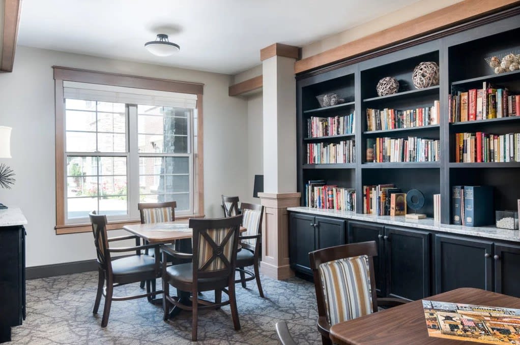 A cozy library reading room featuring bookshelves filled with books, a wooden table, and chairs by large windows.