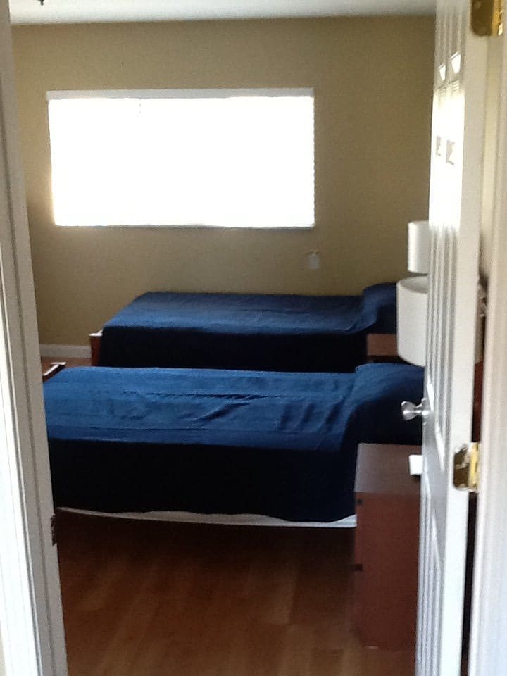 A minimalist bedroom featuring two twin beds with blue covers, a window, and basic furnishings.