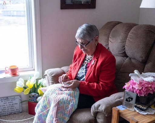 An elderly woman enjoys a cozy afternoon knitting in her comfortable chair, surrounded by vibrant flowers.