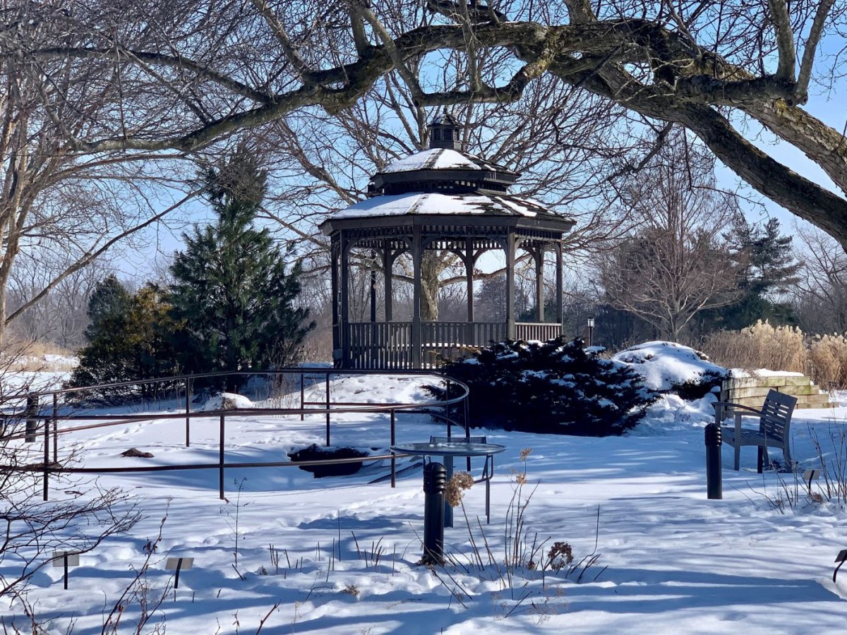 A picturesque gazebo stands amid a snowy winter landscape, surrounded by trees and enclosed by a fence.