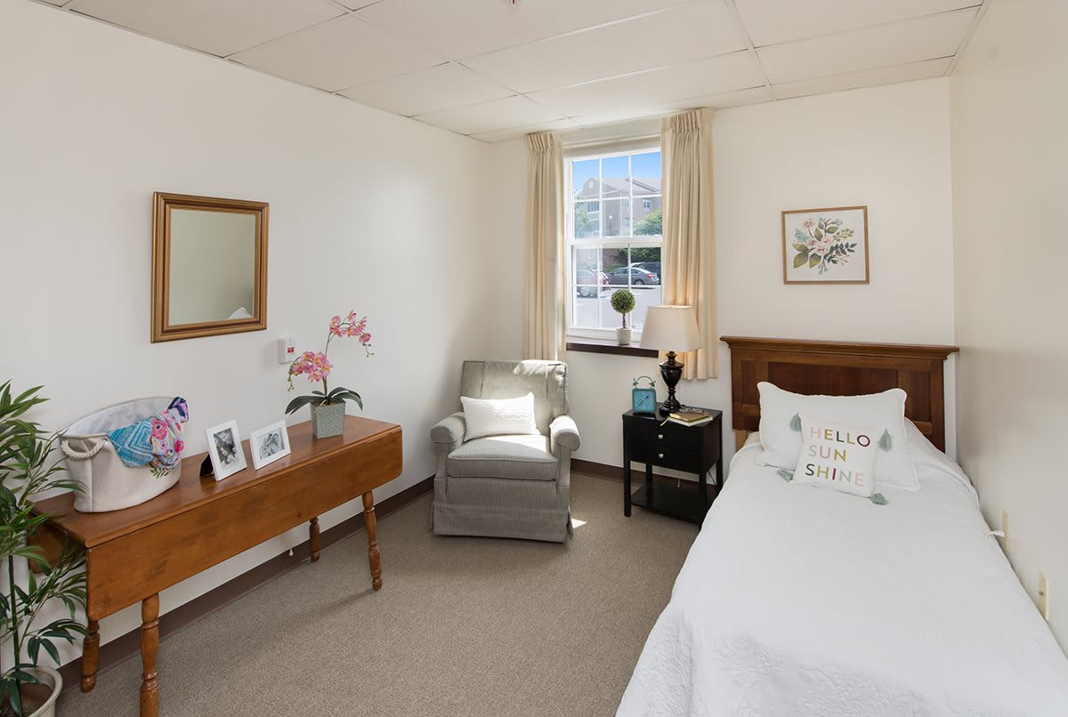 A cozy bedroom with a bed, armchair, and decorated dresser near a window.