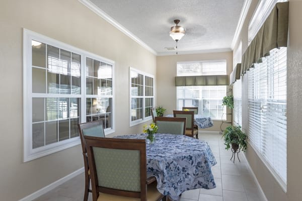 A bright sunroom dining area with tables and chairs, decorated with floral tablecloths and large windows allowing natural light.