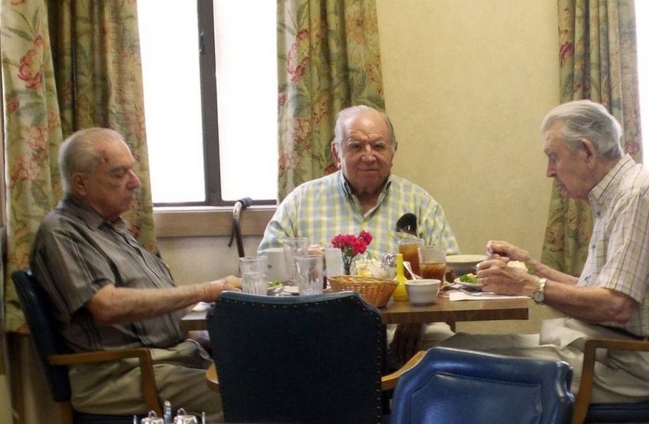 Three elderly individuals are seated around a dining table, enjoying a meal together in an indoor setting.