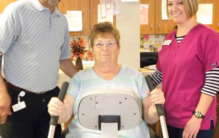 A woman using exercise equipment with the assistance of two therapists.