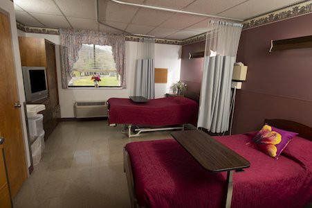 A hospital room with two beds separated by curtains, featuring maroon bedspreads and a window view.