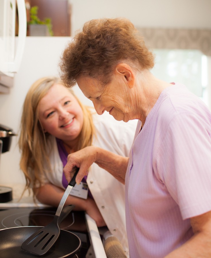 A caregiver assists an elderly woman with cooking in the kitchen.