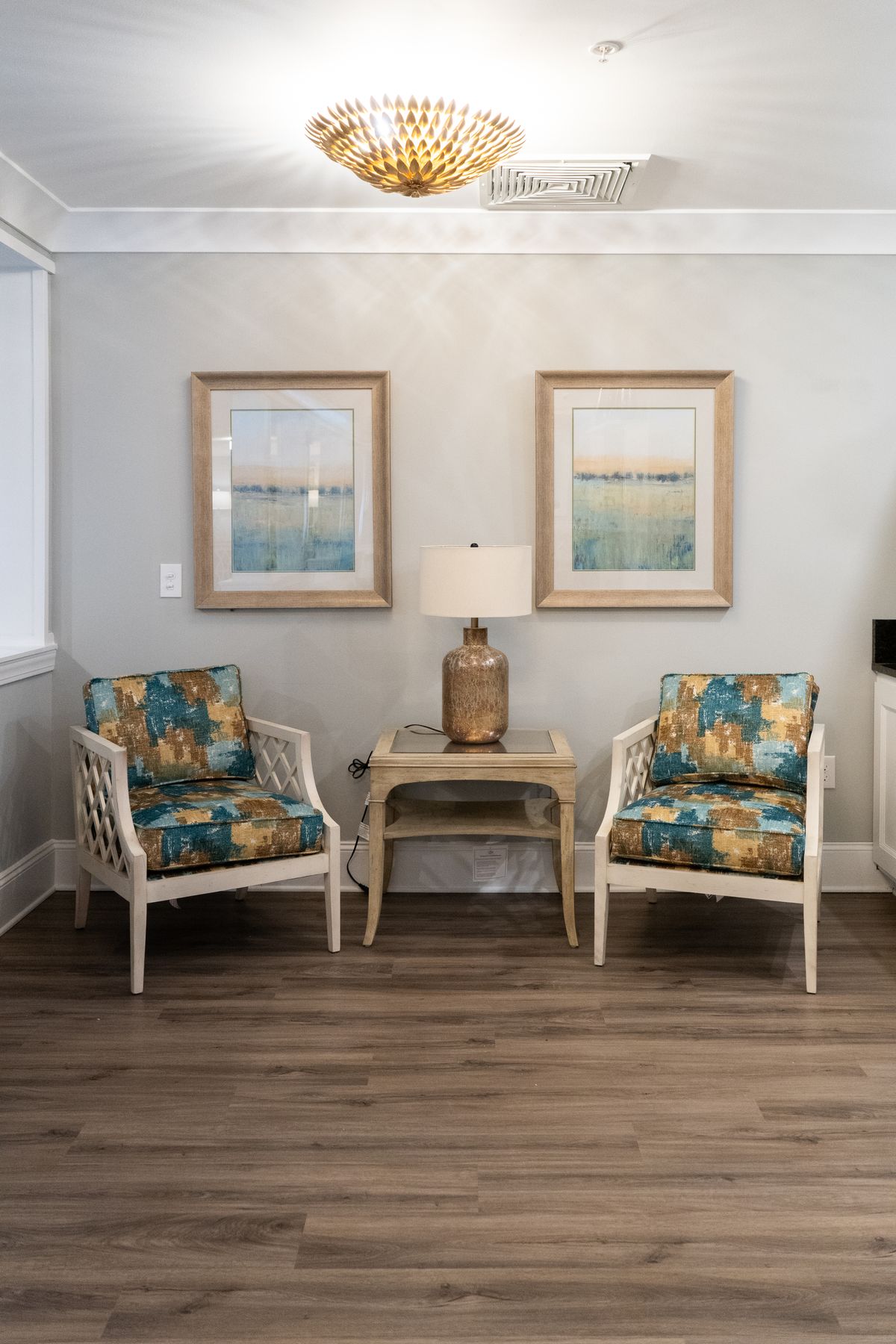 A cozy living room setup with two patterned armchairs, a lamp on a wooden side table, and framed artwork on the wall.