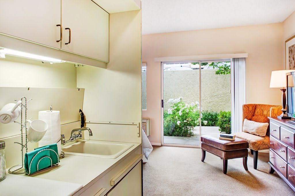 A cozy interior view of a small kitchen area leading to a bright living space with large windows and greenery outside.