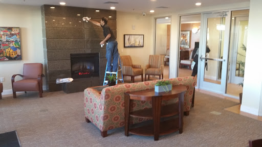 A man is painting a wall in a cozy living room with a fireplace, surrounded by comfortable seating.