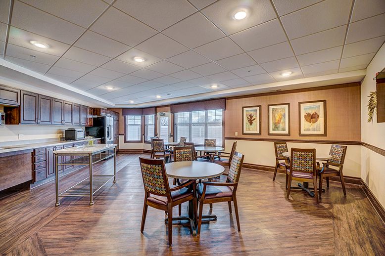 A spacious dining area with multiple tables and chairs, featuring a kitchen area in the background and decorative paintings on the walls.