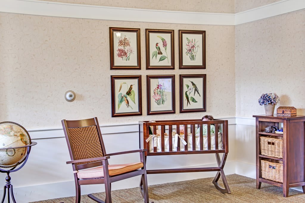 A cozy nursery room with a wooden cradle and rocking chair, adorned with framed artworks on the wall.