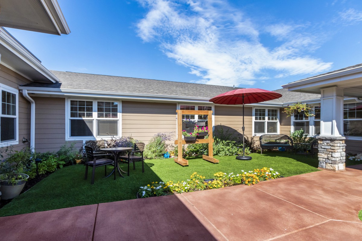 A cozy garden patio with outdoor seating, a swing set, and colorful flowers under a clear blue sky.
